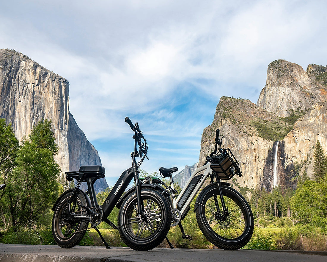 Two electric bikes parked side by side on pavement with scenic mountain cliffs and blue sky in the background.”